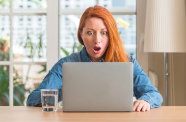 Redhead woman using computer laptop at home scared in shock with a surprise face, afraid and excited with fear expression